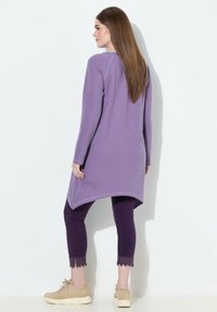 Fioletowa, długa tunika z długim rękawem i zakrzywionym dołem, w połączeniu z ciemnofioletowymi legginsami ozdobionymi koronkowymi detalami oraz beżowymi sneakersami na masywnej podeszwie.