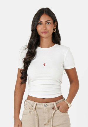 Witte t-shirt met korte mouwen en een aansluitend ontwerp, met een kleine rode graphic op de voorkant. Gecombineerd met beige broek en een horloge.