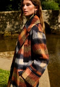 Manteau en tartan en laine présentant de vives motifs orange, marine et crème. Les détails remarquables comprennent un col cranté et des poches latérales.