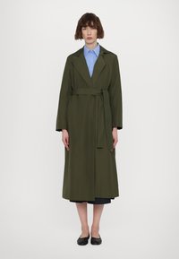 Olijfgroene trenchcoat met een strikceintuur, lange mouwen en een vloeiende uitstraling. Draagt over een lichtblauw overhemd, gecombineerd met zwarte flats.