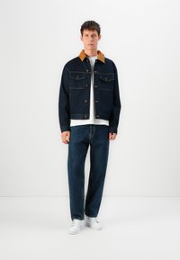 Donker spijkerjack met een bruine corduroy kraag, twee borstzakken en knoopsluiting. Gecombineerd met losvallende donkere jeans en witte sneakers.