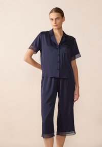 Jūras spēku zīda pidžamu komplekts ar īsām piedurknēm pogājamu topu ar mežģīņu apdari un ceļgala garuma biksēm ar saskaņojošām mežģīņu akcentiem pie apakšmalas.