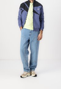 Casual outfit featuring a blue and black zip-up jacket, lime green t-shirt, light blue jeans, and beige sneakers with black accents.