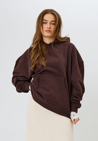 Sudadera marrón de gran tamaño en tejido suave, con un bolsillo frontal, corte relajado y capucha ajustable con cordones, llevada sobre una falda beige clara.