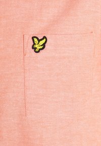 Lichtroze shirt met korte mouwen en een voorzak voorzien van een zwart-gele geborduurde adelaar. Gladde stoftextuur.