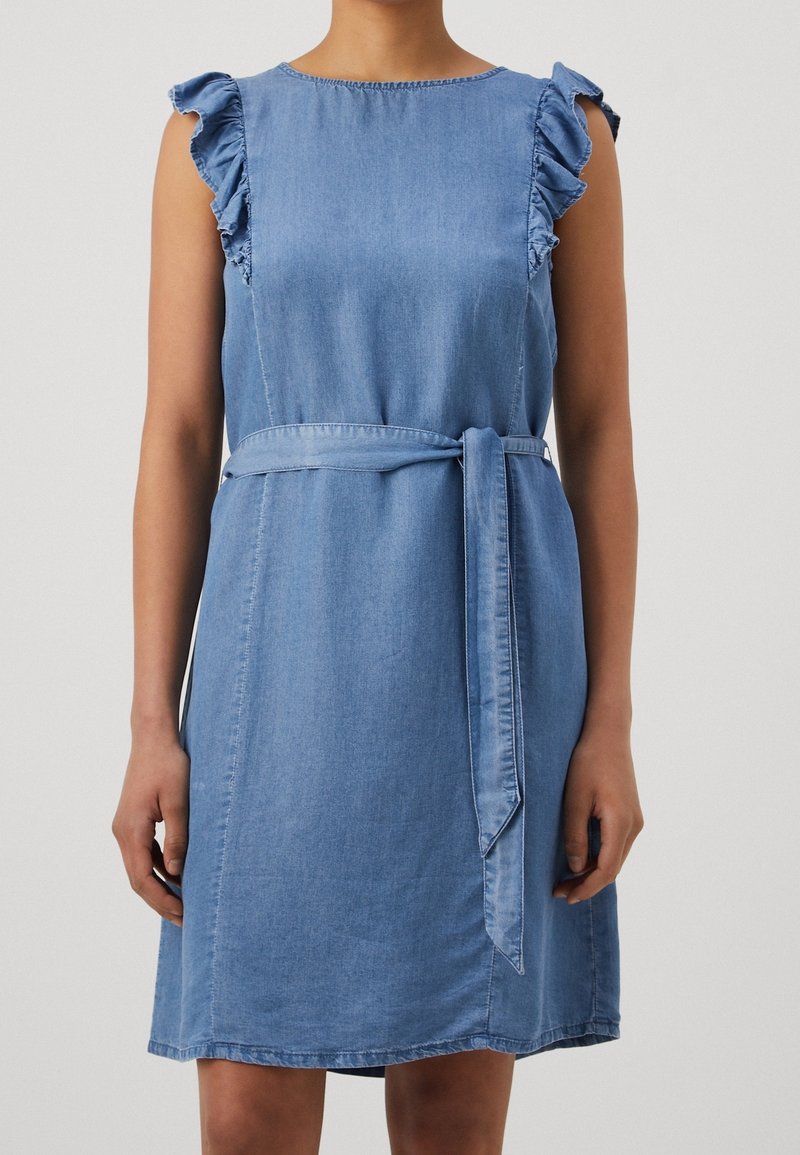 Femme portant une robe en denim bleu clair sans manches avec des détails froncés aux épaules et une ceinture nouée à la taille.