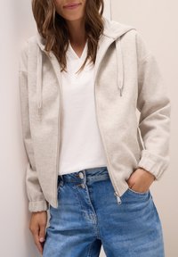 Haut de sweatshirt zippé gris clair avec une texture douce, cordons réglables et poches latérales, porté par-dessus un t-shirt blanc à col en V et un jean bleu.