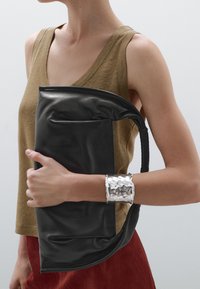 Zwarte leren handtas met een gladde afwerking, gebogen bovenrand en een zijvak. Het model draagt de tas onder haar arm en heeft een beige mouwloos topje aan.