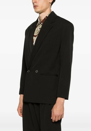 Uomo che indossa un blazer nero doppiopetto sopra una camicia fantasia, in piedi davanti a uno sfondo bianco uniforme, rivolto leggermente a sinistra.