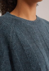 Suéter de punto en color azul verdoso, con un patrón de relieve texturizado. La tela tiene un aspecto suave y difuso, con un diseño clásico de cuello redondo.