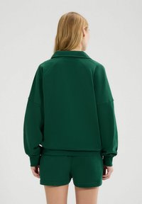 Sudadera verde con cuello, de corte oversize y puños elásticos, combinada con pantalones cortos a juego. Texture suave y detalles de diseño mínimos.