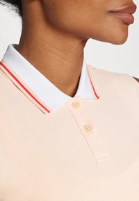 Polo tričko z jemného broskvového materiálu s bílým síťovým límcem a červenými pruhy, se třemi knoflíky a hladkou strukturou. Detailní pohled.