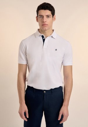 Camisa polo blanca de manga corta con un cuello azul marino, con un pequeño emblema en el pecho. Combinada con pantalones oscuros azul marino.