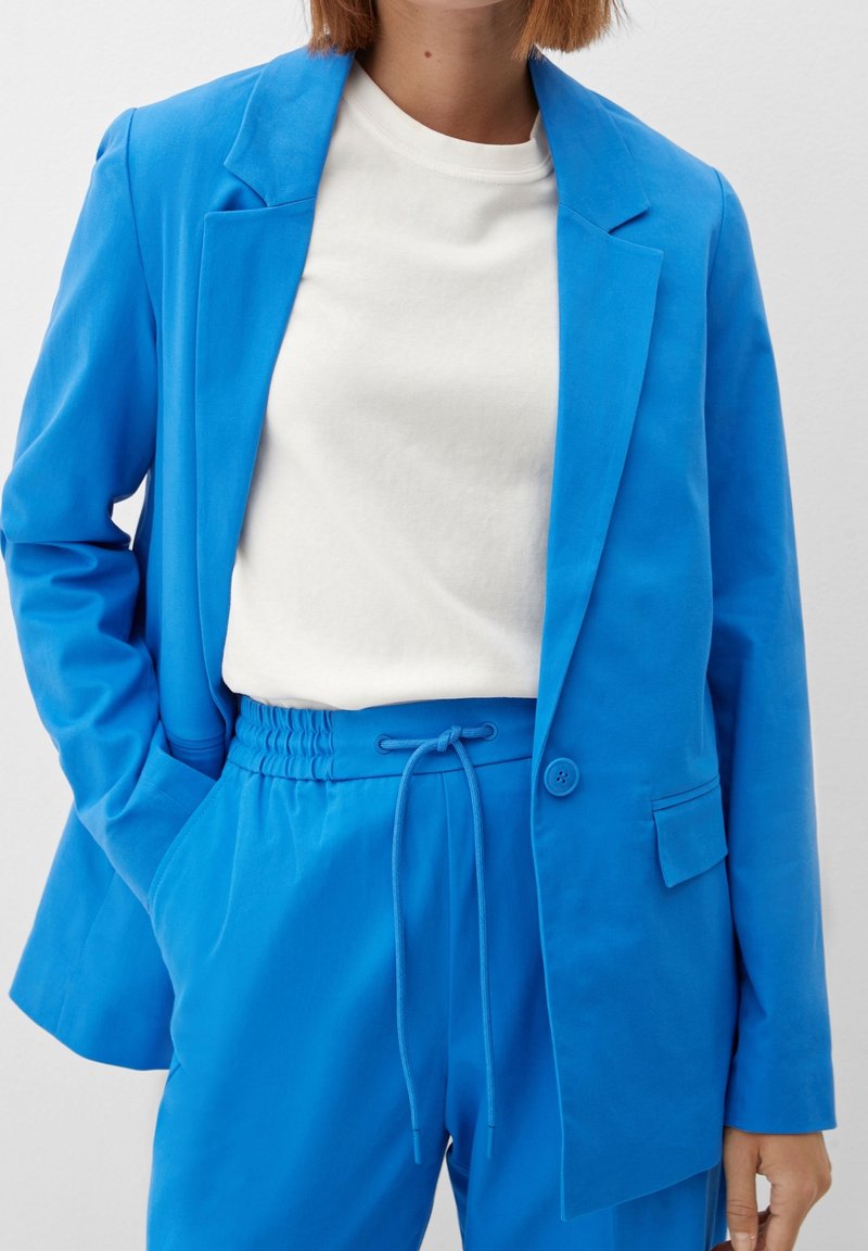 Femme portant un blazer bleu vif et un pantalon à cordon assorti avec un t-shirt blanc à col rond, main dans la poche, sur un fond uni.