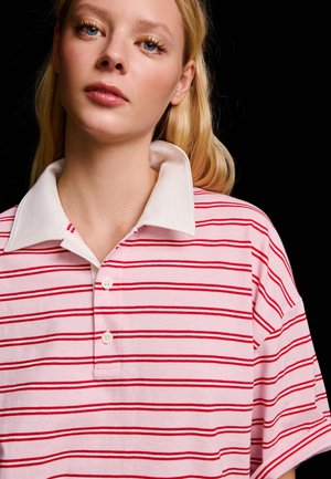 Mladá žena s blond vlasmi, oblečená v ružovom rugby tričku s červenými horizontálnymi pruhmi a bielym golierom, na čiernom pozadí.