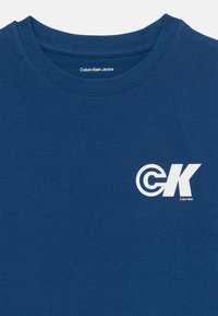 Tamsiai mėlynas medvilnės marškinėliai su apvalia iškirpte, puošti balta "CK" logotipu ir tekstu "Calvin Klein Jeans" apatiniame dešiniajame kampe.