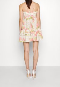 Trägerloses Blumenkleid mit rüschengeschmücktem Saum, das pinke und grüne Muster auf einem hellen Hintergrund zeigt; getragen mit beigen, offenen High Heels.