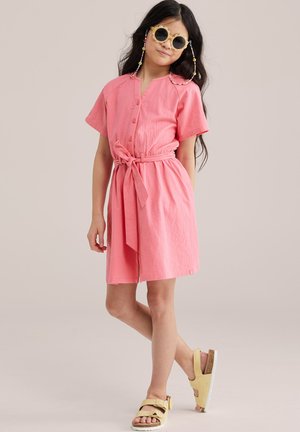 Ragazza in piedi con una gamba incrociata, che indossa un vestito rosa con cintura, sandali gialli, occhiali da sole rotondi e catene per occhiali di perline.