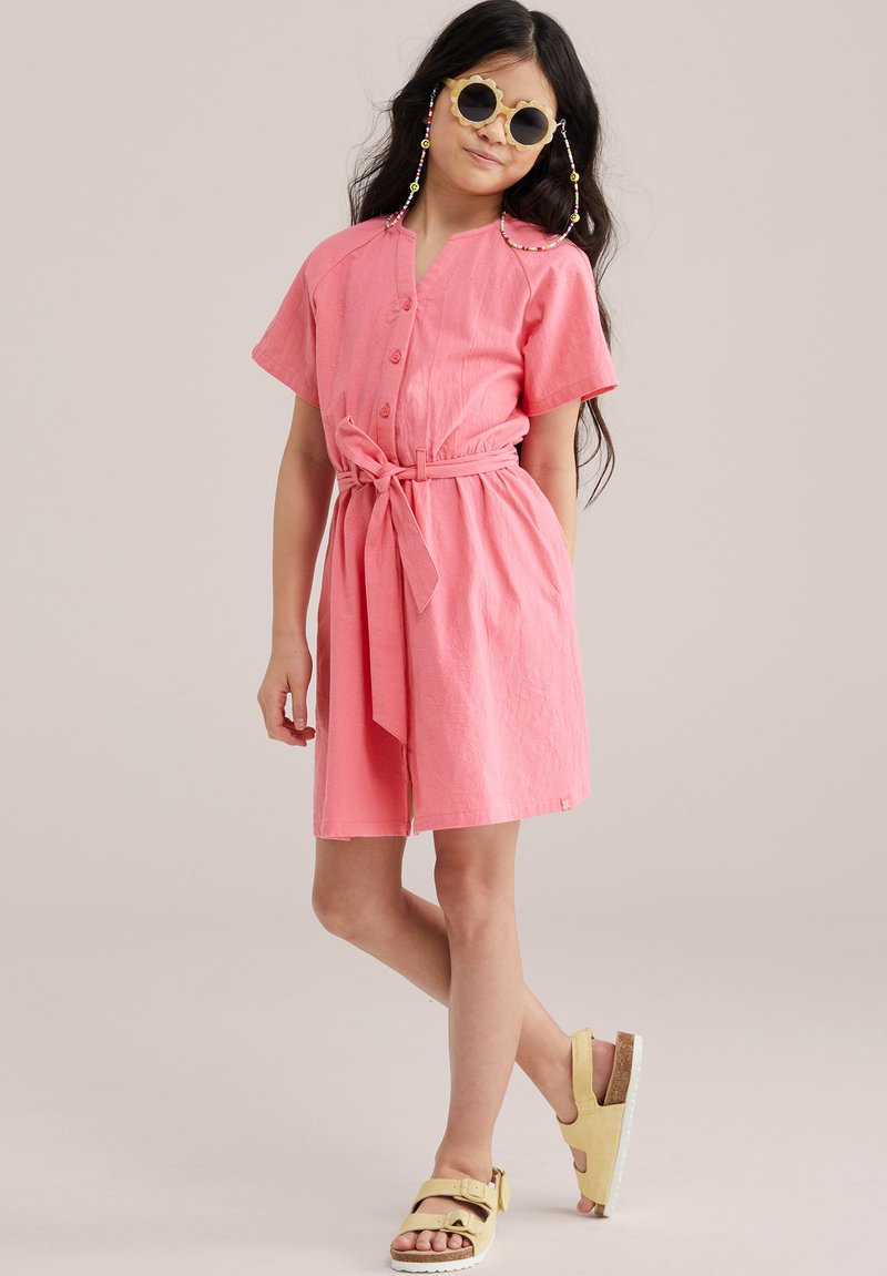Mädchen steht mit einem Bein über dem anderen, trägt ein rosa Kleid mit Gürtel, gelbe Sandalen, runde Sonnenbrille und Perlenbrillenketten.