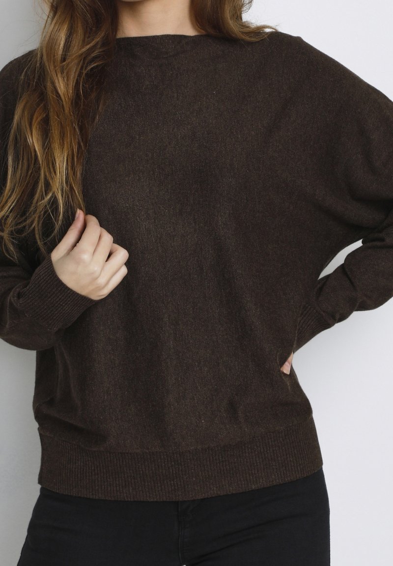 Femme portant un pull tricoté de couleur brun foncé à manches longues, tenant un côté près du col, debout devant un fond uni.