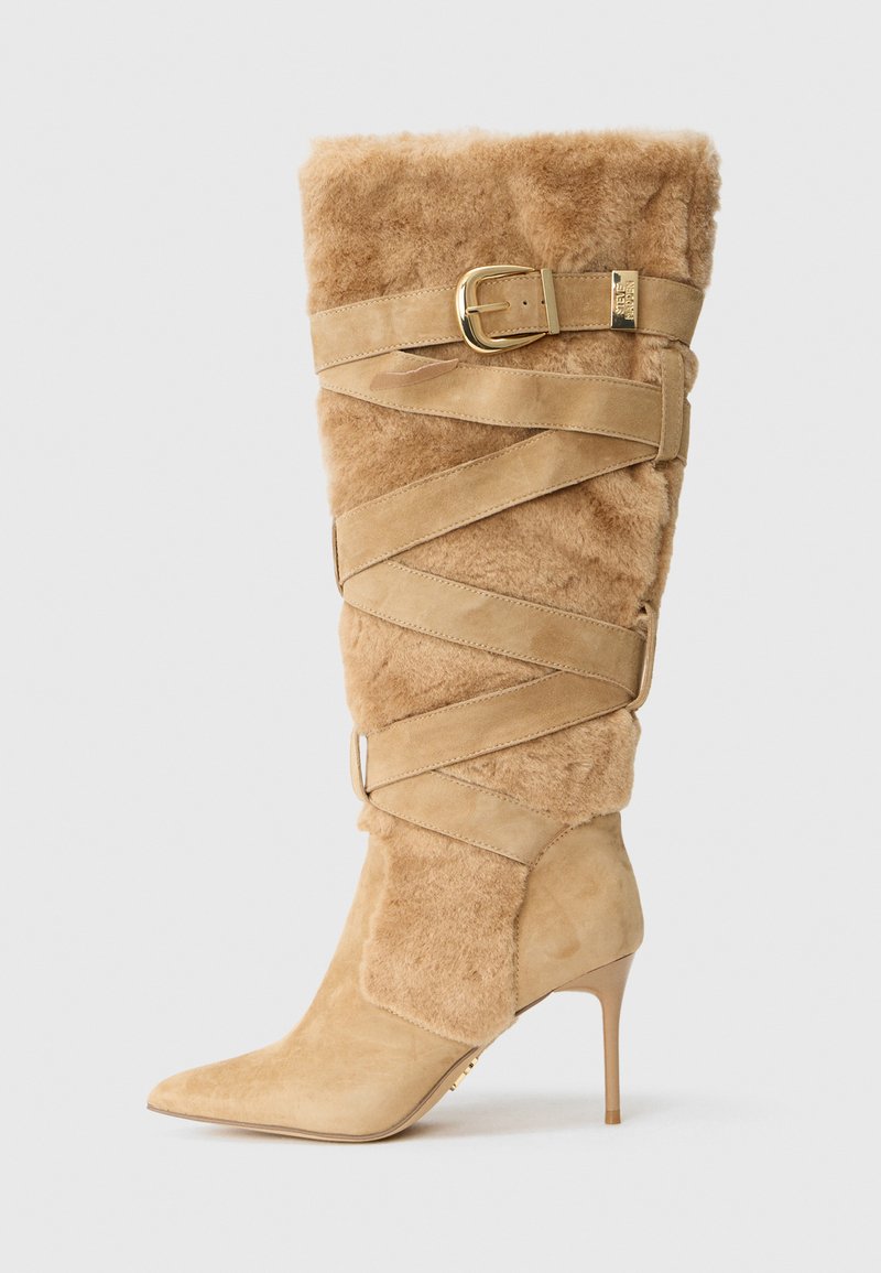 Botă bej din suede până la genunchi, cu toc stiletto și vârf ascuțit, textură pufoasă din blană și curea încrucișată care se înfășoară cu cataramă aurie pe lateral.