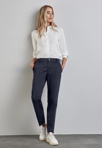 Nainen, jolla on valkoinen napillinen paita, tummansiniset housut ja valkoiset tennarit, seisoo käsineen taskuissa yksinkertaista seinää vasten.