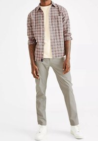 Geruit button-up shirt in rood, beige en paarse tinten, gedragen over een crèmekleurig T-shirt, gecombineerd met lichtgrijze broek en witte sneakers.