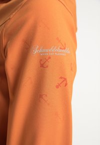 Oranžová látka s hladkým povrchem, se vzorem kotev v mírně tmavším odstínu a logem v bílém písmu na rameni.