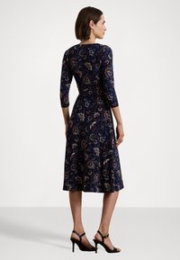Abito floreale blu navy con maniche a tre quarti, corpina aderente, gonna a campana e motivi intricati in oro, rosso e bianco. Sandali neri con tacco alto.