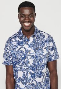Camicia a maniche corte con bottoni in bianco e blu navy con motivo floreale, dotata di taschino sul petto e colletto rotondo. Tessuto leggero.