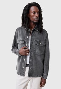 Chaqueta de mezclilla gris con dos bolsillos en el pecho, cierre de botones y un corte holgado. Presenta una textura sutil y botones plateados en contraste.