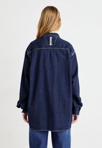 Mørkeblå denimskjorte med afslappet pasform, knappede ærmer og et synligt bagetiket. Har kontraststing og en klassisk krave.