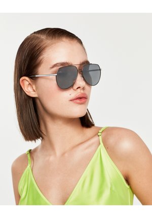 Fată tânără cu părul scurt, castaniu, purtând ochelari de soare aviator gri mari și un maiou satinat verde lime, privind spre dreapta.