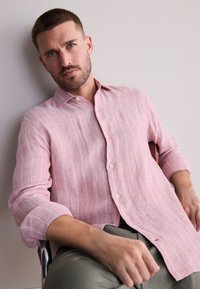 Ljusrosa linneskjorta med vita vertikala ränder, knäppt med en knapp och uppkavlade ärmar, bärs över olivgröna byxor.