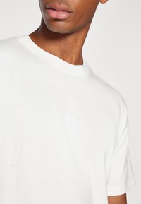 Hvit bomulls t-skjorte med rund hals og korte ermer. Merkbar detalj: liten hvit brodert logo på brystet. Glatt tekstur.