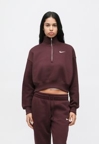 Hanorac burgund de tip cropped, cu guler înalt și fermoar pe jumătate, având un mic logo alb Nike pe piept, asortat cu pantaloni potriviți.