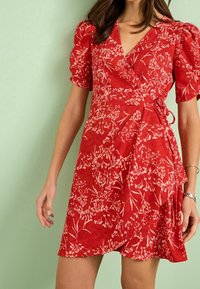 Robe portefeuille rouge avec manches bouffantes, motif floral blanc et détail de nouage sur le côté. Confectionnée dans un tissu texturé, avec une silhouette fluide.