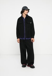 Zwarte textuurjack over een paars geruit shirt, gecombineerd met losse zwarte broek en zwarte sneakers met rode accenten, evenals een grijze pet.