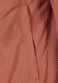 Gros plan d'une poche de veste de couleur rouille avec des coutures visibles et une doublure en tissu texturé sur le côté droit.
