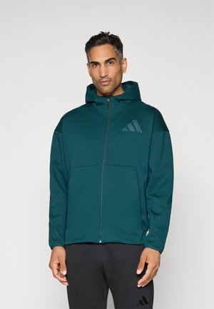 Hombre con chaqueta con cremallera color teal y capucha, y pantalones negros, de pie contra un fondo claro y liso.