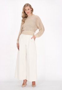 Beżowy sweter z dzianiny o otwartym splocie, sparowany z białymi spodniami o szerokich nogawkach z przodu z plisami i kieszeniami bocznymi. Schludny, minimalistyczny design.