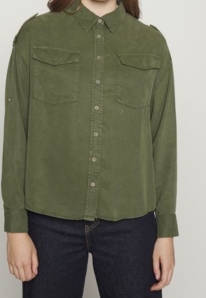 Nainen, jolla on yllään oliivinvihreä nappirapaita, jossa kaksi rintataskua, ja tummansiniset farkut, seisoo yksinkertaisen vaalean taustan edessä.