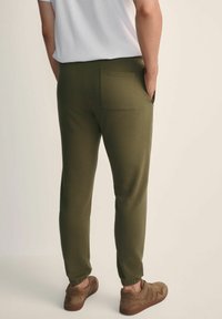 Olive zielone spodnie dresowe wykonane z miękkiego materiału, z pojedynczą kieszenią z tyłu, elastycznymi mankietami i luźnym krojem. Brązowe buty dopełniają wygląd.