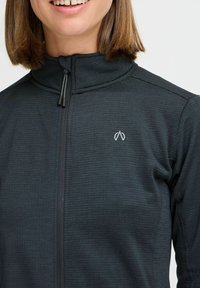 Melna jaka ar rāvējslēdzēju un augstu apkakli, teksturēta auduma, ar mazu logotipu kreisajā pusē. Aprīkota ar divām melnām rāvējslēdzēja rokturiem.