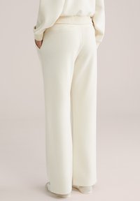 Person iført cremegule bukser med vide ben og matchende top, hænderne i lommerne, stående på en lys baggrund.