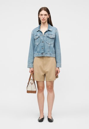 Tânără purtând o geacă de denim de culoare bleu deschis, pantaloni scurți bej până la genunchi, balerini negri și ținând o geantă bej cu elemente maro.