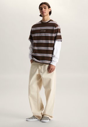 Joven de pie con las manos en los bolsillos, vistiendo una camiseta marrón con rayas de Vans sobre una camiseta blanca de manga larga, pantalones anchos color crema y zapatos slip-on a cuadros.