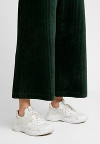 Witte sneakers met dikke zolen en veters, gecombineerd met wijduitlopende donker groene corduroybroek met een verticale geribbelde textuur.