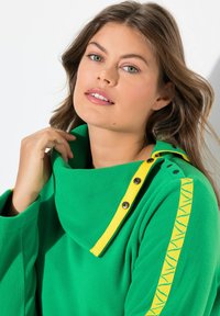 Sudadera de felpa verde con un amplio cuello, cinta de acento amarilla y detalles de botones. Presenta un patrón de rayas texturizado en la manga.