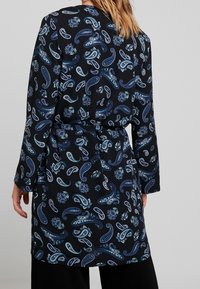 Lång svart kappa med paisley- och blommönster i blått och teal. Har ett bälte och långa ärmar, tillverkad av ett mjukt tyg.